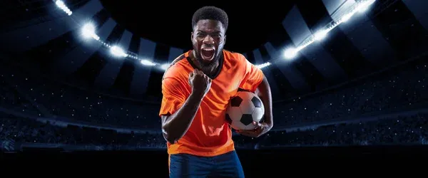 Victory. Excited professional soccer, football player in football kit standing with ball and shouting over dark night stadium with flashlights background. — Stock Photo, Image